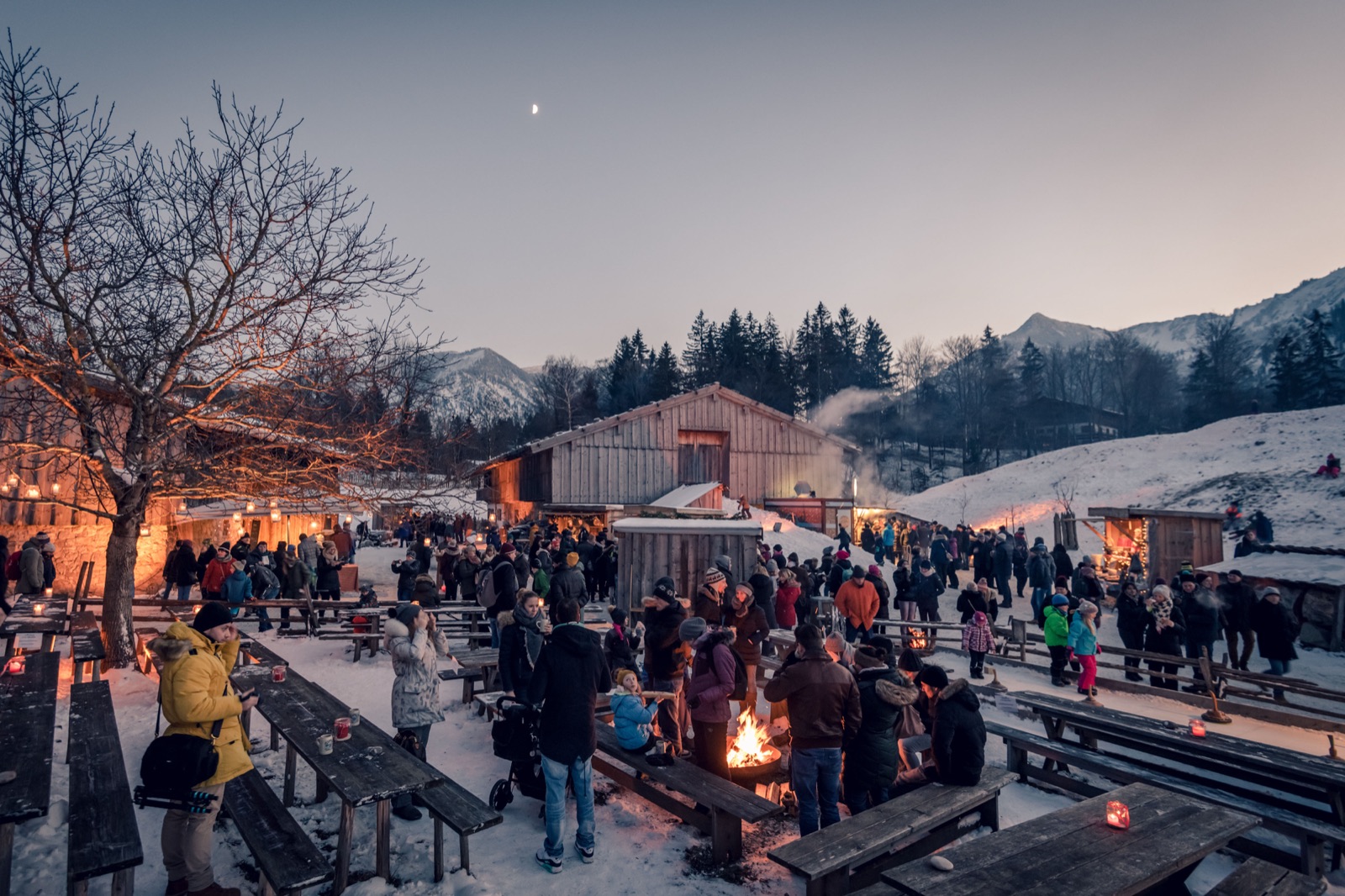 Schliersee im Winter: Besucher am Lagerfeuer vor schneebedeckten Almhütten und Bergen in der Dämmerung