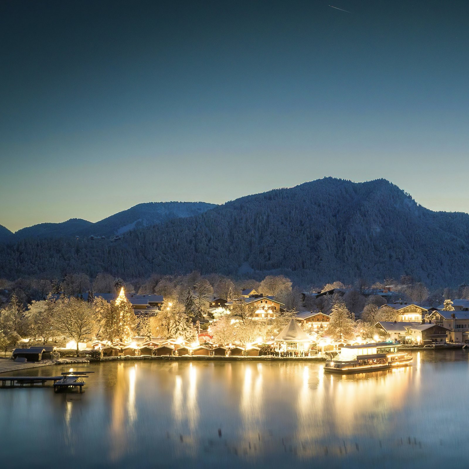 Tegernsee zur blauen Stunde: beleuchteter Weihnachtsmarkt und Schiff am Seeufer, gespiegelt im Wasser vor winterlicher Bergkulisse